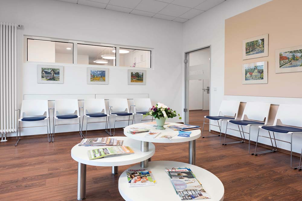 Wartezimmer mit Stuhlreihen auf zwei Seiten, Glastür und im Zentrum einen dreigeteilten Tisch in verschiedenen Ebenen.